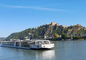 Statek wycieczkowy Viking Kara, płynący po Renie W tle wznosi się monumentalna twierdza Ehrenbreitstein w niemieckim mieście Koblenz. Forteca usytuowana jest na wysokim, skalistym wzgórzu porośniętym zielenią.
