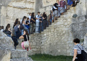 Uczestnicy wycieczki wspinają się po kamiennych schodach przy starych murach na część zabytkowej budowli wykutej w skale.