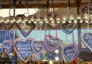 Na zdjęciu widać stoisko z jarmarku, na którym wiszą liczne pierniki w kształcie serc. Są zawieszone na wstążkach i ułożone w kilku rzędach. Na piernikach znajdują się niemieckie napisy, m.in. wyrażenia sympatii i miłości. W tle widać elementy stoiska oraz refleksy świateł, co nadaje zdjęciu ciepły, jarmarczny klimat. Liliana Zdjęcie 1 Na zdjęciu widać grupę dorosłych osób uczestniczących w kursie doskonalącym. Uczestnicy stoją i siedzą w sali szkoleniowej, uśmiechają się i pokazują gest serca dłońmi. Jedna z osób trzyma worek z drobnymi prezentami, co podkreśla ciepłą, życzliwą atmosferę. Zdjęcie oddaje integrację grupy i pozytywny klimat wspólnej nauki. Liliana Zdjęcie 2 Na zdjęciu widać diabelskie koło w Lubece nocą, rozświetlone kolorowymi światłami. Konstrukcja tworzy efektowny, wielobarwny krąg na tle ciemnego nieba. W tle znajdują się zabytkowe kamienice, które podkreślają świąteczno-miejski klimat miejsca.