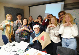 Na zdjęciu widać grupę dorosłych osób uczestniczących w kursie doskonalącym. Uczestnicy stoją i siedzą w sali szkoleniowej, uśmiechają się i pokazują gest serca dłońmi. Jedna z osób trzyma worek z drobnymi prezentami, co podkreśla ciepłą, życzliwą atmosferę. Zdjęcie oddaje integrację grupy i pozytywny klimat wspólnej nauki.
