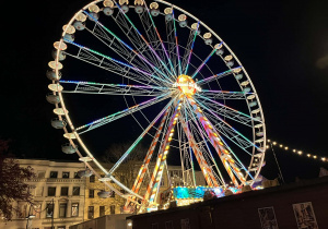 Na zdjęciu widać diabelskie koło w Lubece nocą, rozświetlone kolorowymi światłami. Konstrukcja tworzy efektowny, wielobarwny krąg na tle ciemnego nieba. W tle znajdują się zabytkowe kamienice, które podkreślają świąteczno-miejski klimat miejsca.