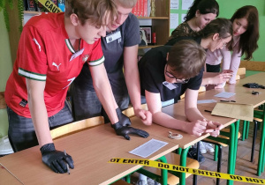 Uczniowie, pracując w grupach przy stanowisku oznaczonym taśmą „Do not enter”, analizują wskazówki i rozwiązują zadania w trakcie detektywistycznej gry.