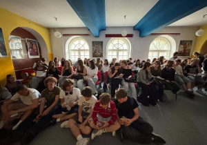 Grupa uczniów siedzi na krzesłach i na podłodze, trzymając kartki oraz telefony podczas przygotowań do quizu.