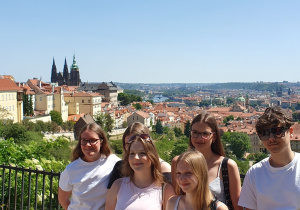 Grupa uczniów pozuje do zdjęcia na punkcie widokowym, a w tle rozciąga się panorama Pragi z charakterystycznymi czerwonymi dachami i wieżami.