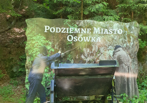 Tablica „Podziemne Miasto Osówka” ustawiona przy torach i wagoniku górniczym.