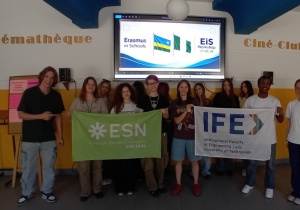 Grupa uczestników warsztatów pozuje w szkolnej sali, trzymając banery ESN i IFE na tle prezentacji „Erasmus in Schools”.