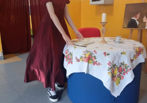 Uczennica w stylizowanym stroju z epoki odgrywa scenę z twórczości Jane Austen przy starannie przygotowanej scenografii, nawiązującej do realiów XIX wieku.