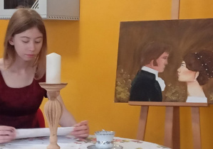 Uczennica przy stole odczytuje fragment tekstu w scenie inspirowanej twórczością Jane Austen, na tle stylizowanej dekoracji z portretem bohaterów.