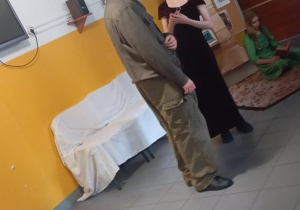 Dwie osoby w kostiumach z epoki odgrywają dialog, a w tle widoczna jest scenografia i uczennica czytająca książkę.