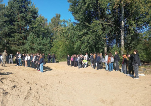 Uczniowie klas pierwszych zgromadzeni na leśnej polanie wysłuchują instrukcji przed rozpoczęciem kolejnych zadań